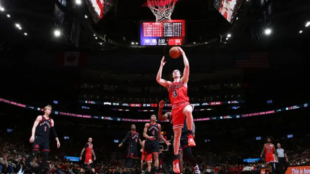Los Angeles clipper player in red uniform goes to put the ball into the net as Raptors players observe. Wide shot shows the stadium in the background with the crowd and promotional screens in the background