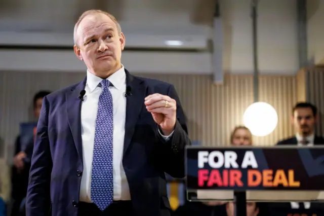Ed Davey in a suit and tie in front of podium saying for a fair deal with blurred people behind