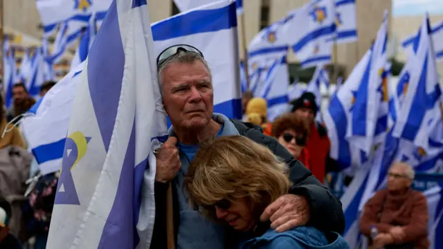 Man holding a flag hugs a crying woman. In the background, there is a sea of Israeli flags and people