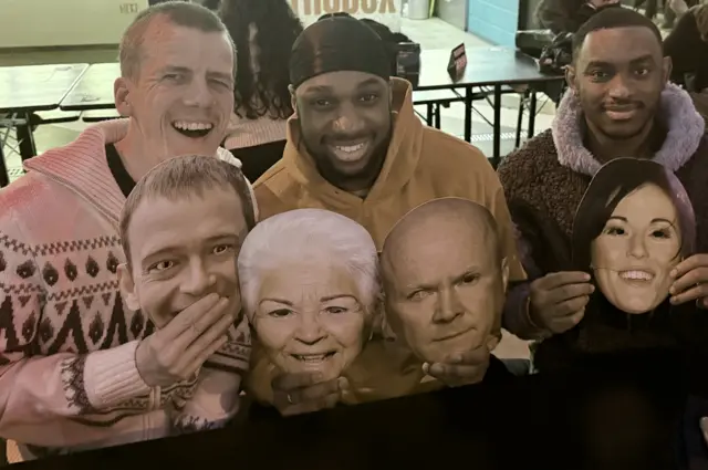 Three men holding cardboard cut-out masks of EastEnders characters.