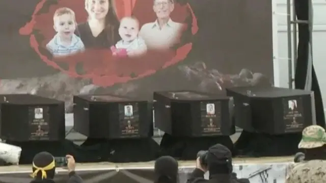 Four black coffins lined up on a stage, with an armed Hamas member stood by