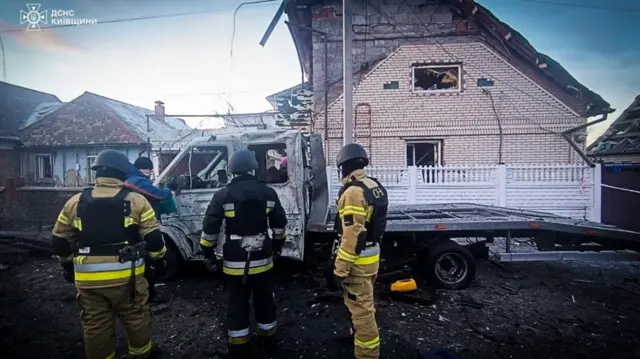 A bombed out house and truck are looked at by the emergency services