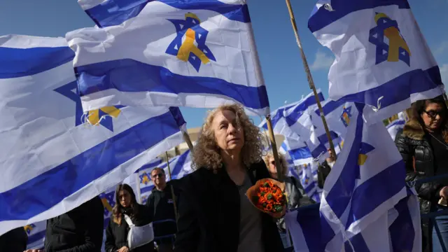 People with flags have gathered at Hostages Square on the day the bodies of four hostages have been returned to Israel. A woman with blonde curly hair and a bouquet of flowers is standing in the centre-front of the image.