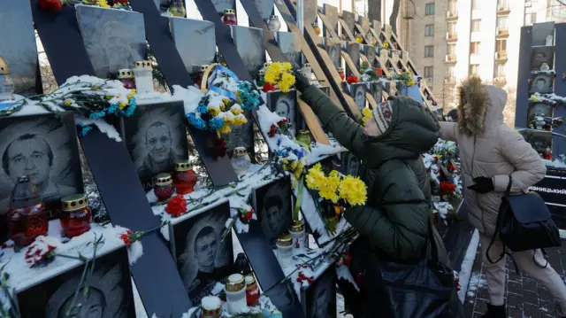 Two women in puffy coats place flowers on memorial showing photos of those who died in the protests.