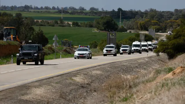 A line of white vehicles drives along a road surrounded by fields