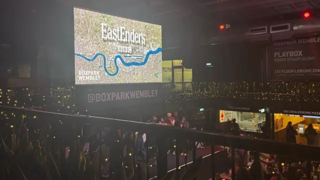 Crowd at Boxpark in Wembley with people stood on stage below EastEnders picture