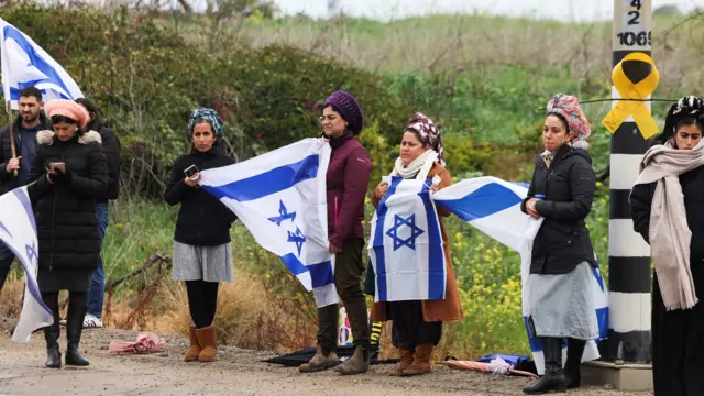 People line a street in Tel Aviv as they wait for bodies to return. There are seven women and a man holding flags