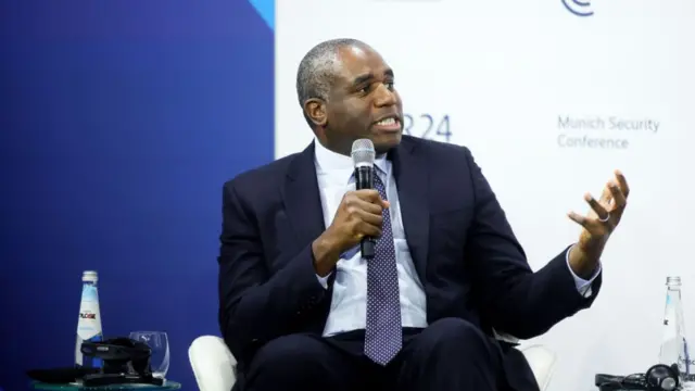 Britain's Foreign Minister David Lammy wears a suit and tie. He sits in front of a backdrop reading Munich Security Conference. He holds a microphone.