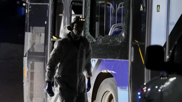 Obscured by shadow, a person in a white forensic suit walks next to the burned shell of a bus