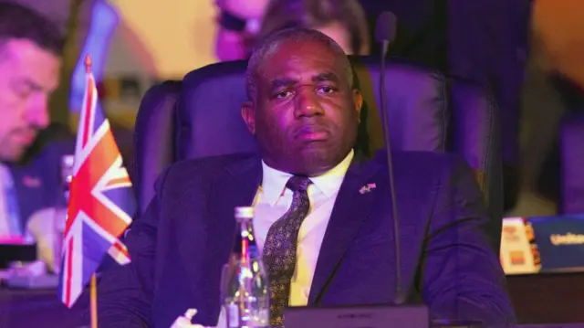 Britain's Foreign Minister David Lammy attends the G20 Foreign Ministers meeting wearing a suit. He sits behind a table with a UK flag on it and a microphone.