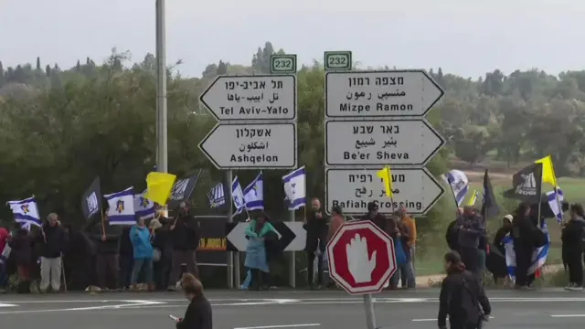 A small crowd stands near a road sign. Many carry Israeli flags, others carry yellow or black flags