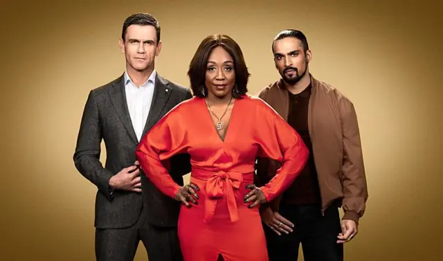 Group portrait of three EastEnders characters, with Denise in the middle wearing a bright orange-red dress, with her hands on her hips and flanked by two men standing behind her The man on the left wears a suit and the one on the right is more casually dressed in a brown sports jacker