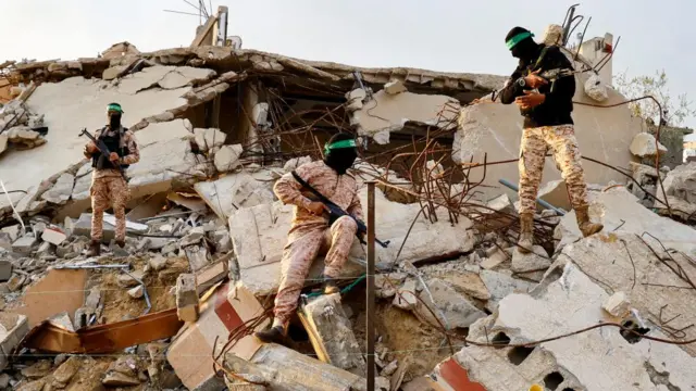 Hamas members in camouflage outfits with black balaclavas and firearms stand on the remnants of a building