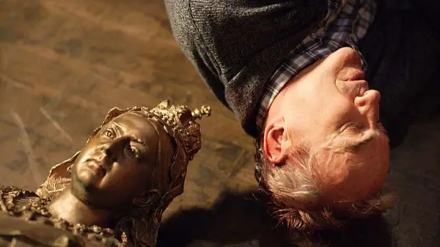 The bust of the Queen Victoria on the floor next to a man who is also lying on the floor with his eyes closed