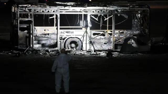 Bus burnt out as man in white suit stands nearby