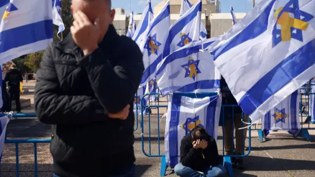 Blue barriers with flags on have been set up in Hostages Square. A man is standing on the left of the image, holding his head, while a woman is sat cross legged in the centre-back of the image with her head in both of her hands.