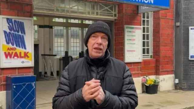 Adam Woodyatt standing in front of the fictional Walford East station on the Eastenders set. He's wearing dark winter clothing and a beanie, clasping his hands together