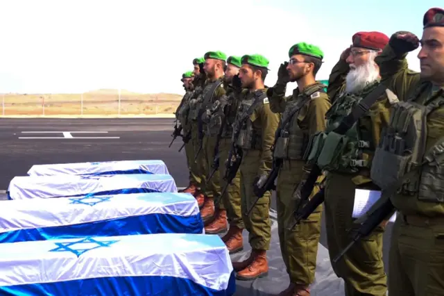 IDF soldiers perform a salute in front of the four lined up coffins which are draped in Israel flags