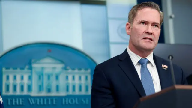 Waltz is on the right of the photo. There is a picture of The White House behind him with those words written underneath as well as the word Washington. Waltz wears a navy suit jacket, striped white shirt and blue tie. He speaks into a small press microphone