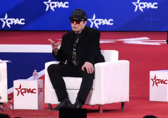 Elon Musk seen on a red, white and blue stage at CPAC. He is wearing all black, including a black hat, and is wearing large reflective sunglasses.