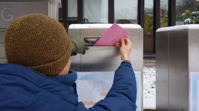 A person in a hat and winter jacket sliding a ballot in an envelope in a post box