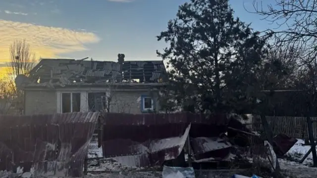 The sun rises behind a damaged house with a broken roof