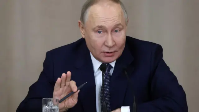 Putin, who has a balding head, wearing a navy suit jacket and tie, speaks as he sits at a wooden table. There's a crystal glass with water in front of him. He has one arm resting on the table, and another held up in front of him as he holds a pen