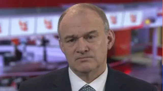 Ed Davey looks serious, he wears a black suit jacket, white shirt and you can just see the top of his patterned tie. He sits in front of a newsroom below, which is blurry but you can see there's screens and computers with desks.