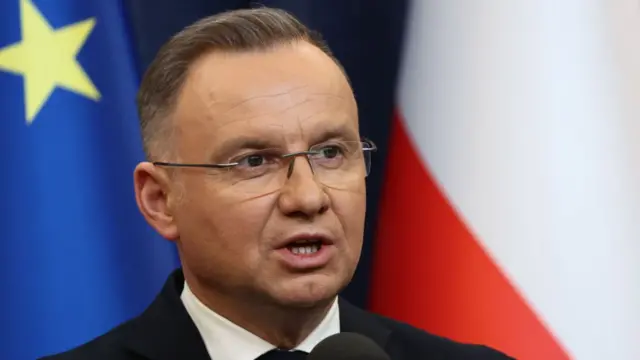 President Duda speaks, and is seen in close up with a flag draped behind him.