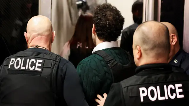Mangioni, wearing a green sweater, white shirt and a bulletproof vest, is escorted out of courtroom by three police officers also wearing bulletproof vests. They are all facing aware from the camera