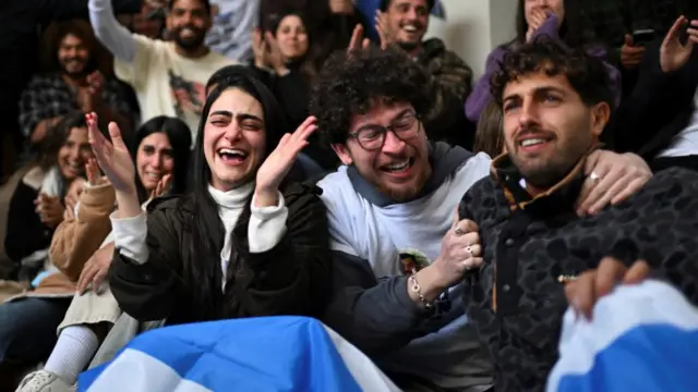 Friends of Omer Wenkert clapping and cheering. Many are seated and some have the Israeli flag on their legs