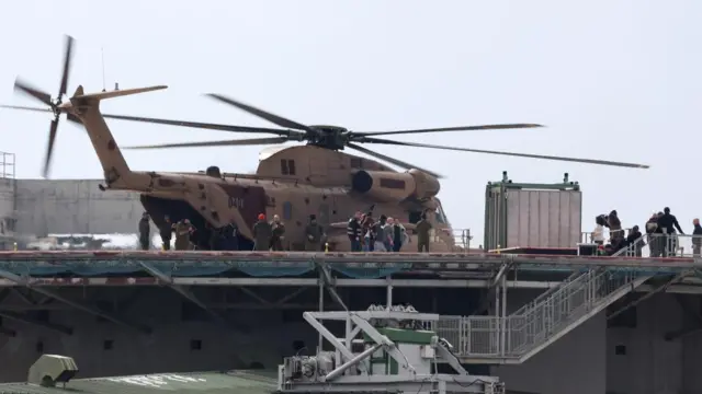 Army helicopter on a platform with group of people stood nearby, with steps leading down to a lower platform.