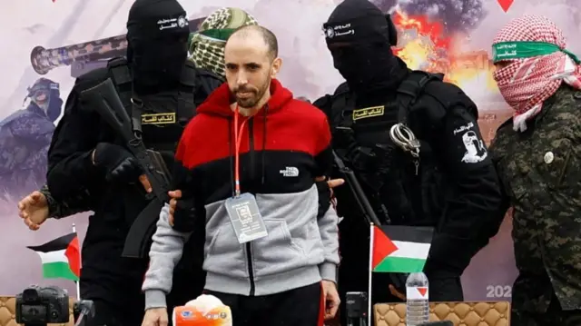 Israeli hostage Tal Shoham on a stage flanked by two armed and masked Hamas members who are holding him by each elbow