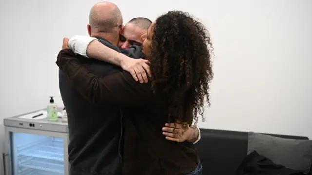 Omer Wenkert hugs two members of his family insider a white-walled room, a dark couch and a grey mini fridge in the background