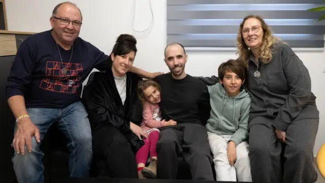 Hostage Tal Shoham sits on a sofa with his family. There are two young children and three adults sat with him, all smiling at the camera.