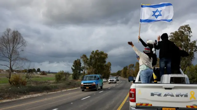 Three people standing on the back of a white car wave an Israeli flag (white background with a blue star of David in the centre, one blue stripe at the top, one matching at the bottom) as a car convoy drives past them. A green field with shrubbery visible to the left of the frame