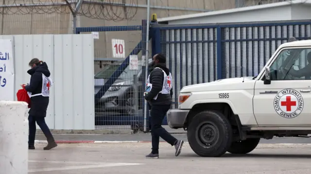 Two members of the International Committee of the Red Cross walk outside Ofer Prison. A woman is walking ahead in a black winter jacket and red cross white vest on; a man is behind her in a black puffer jacket and matching vest, a Red Cross white car behind him