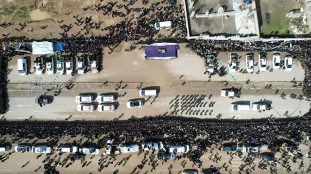 Aerial view of prisoner exchange in Gaza. To the centre left are four Red Cross white cars with a red cross visible at the top. At the centre, is a purple stage where the exchange was filmed, more white cars parked to the top right of the image. Large crowds surround the main scene