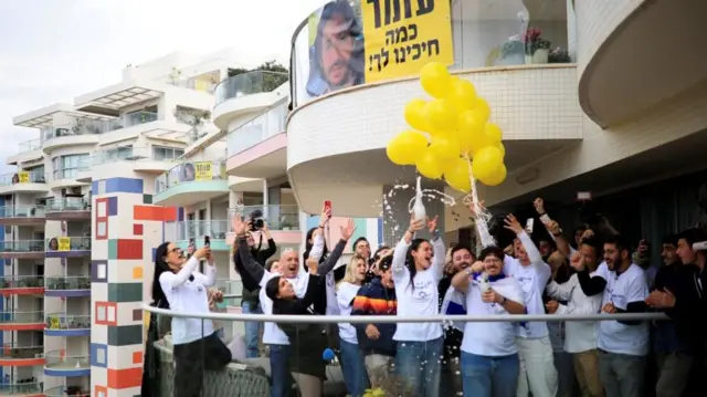 Large group of people cheering on a balcony, with champagne bursting from a bottle, yellow balloons, and a poster of Omer Shem Tov