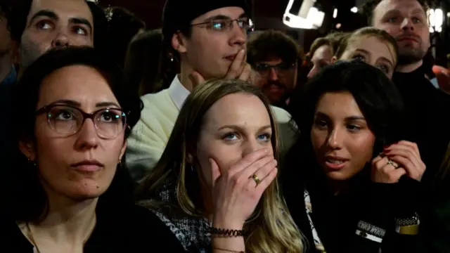 A close up of several people's faces - men and women, looking serious and with their hands over their mouths.