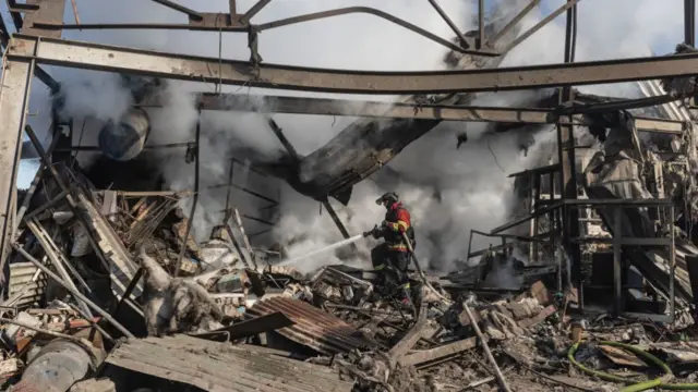 A firefighter standing in a burnt out building with rubble surrounding him. There's smoke around him and he is hosing down the building