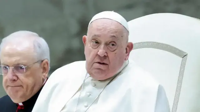 Pope Francis in white robe and hat looking away from camera, sat on white seat with a man in glasses beside him.