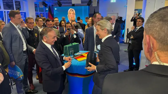 Éric Zemmour giving Alice Weidel a bottle of champagne, surrounded by AfD supporters