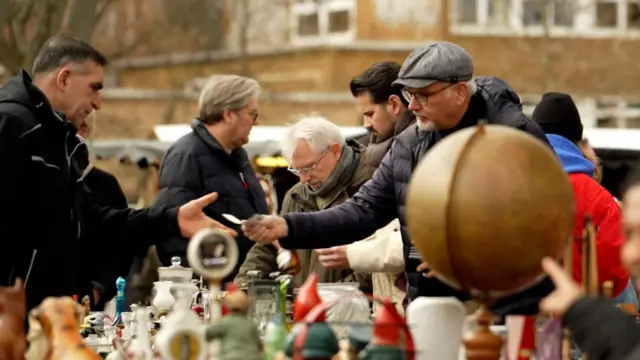 A man wearing a black coat and grey cap hands over cash notes to a stall holder, who is also wearing a black coat, at a market.