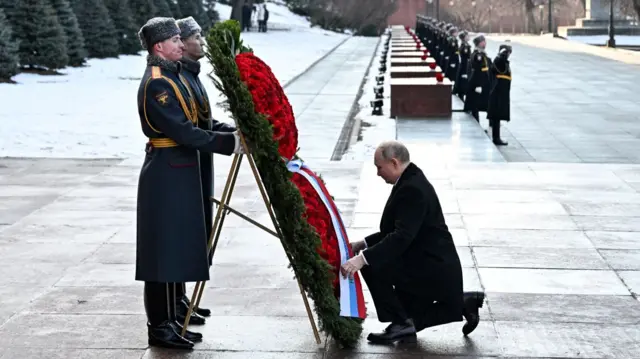 Putin kneeling in front of a large red and green wreath. Behind it are two military personnel in fancy grey and gold jackets. In the background stands a long line of more military men