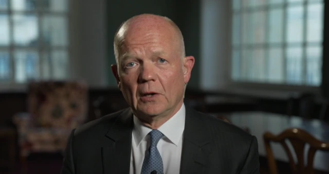 Headshot of Hague who speaks looking into the camera. He wears a black suit jacket, white shirt and blue tie. Behind him is blurry, but it seems there's a table with some chairs around it and an armchair in the room