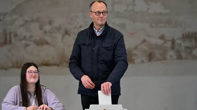 Friedrich Mertz poses for photographers as he casts his ballot into a grey ballot box. He is wearing a blue shirt with a black coat on top. He is wearing black-rimmed glasses.