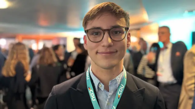 Maximilian smiles directly into the camera. He has short brown hair, brown eyes, and brown-rimmed glasses. He is wearing a blue-striped shirt and black blazer.