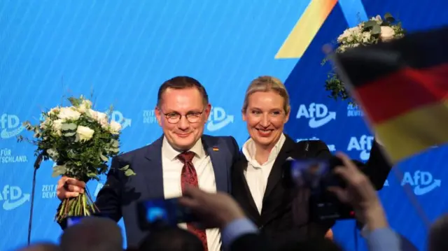 Tino Chrupalla and Alice Weidel holding flowers, arm in arm and smiling.