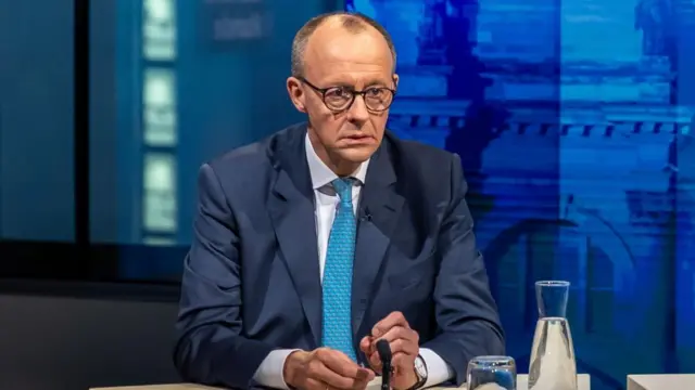 Friedrich Merz on TV wears a blue suit and tie with a white shirt.